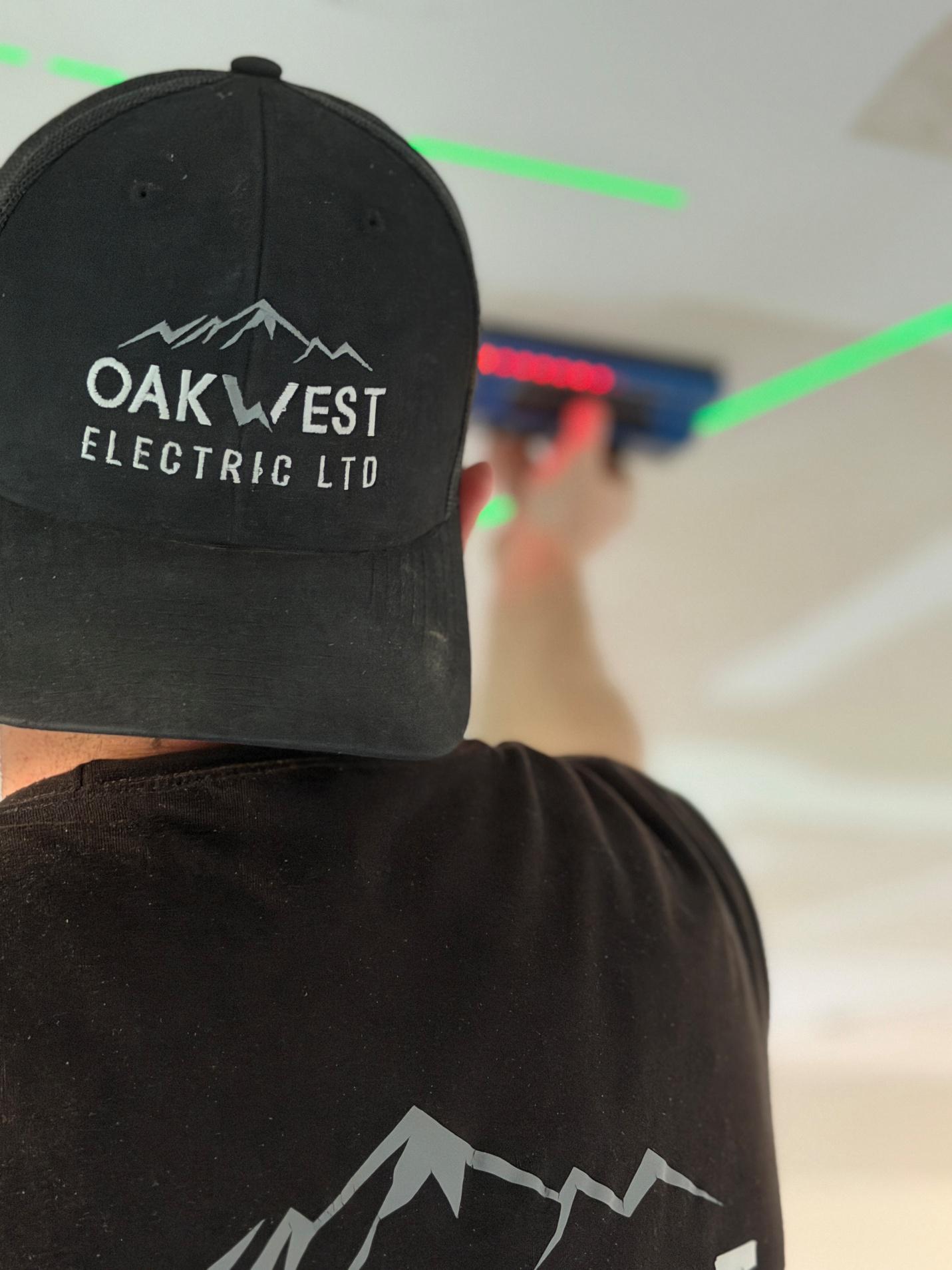 An electrician from Oakwest Electric Ltd using a laser level and professional tools to plan a lighting installation on a ceiling.