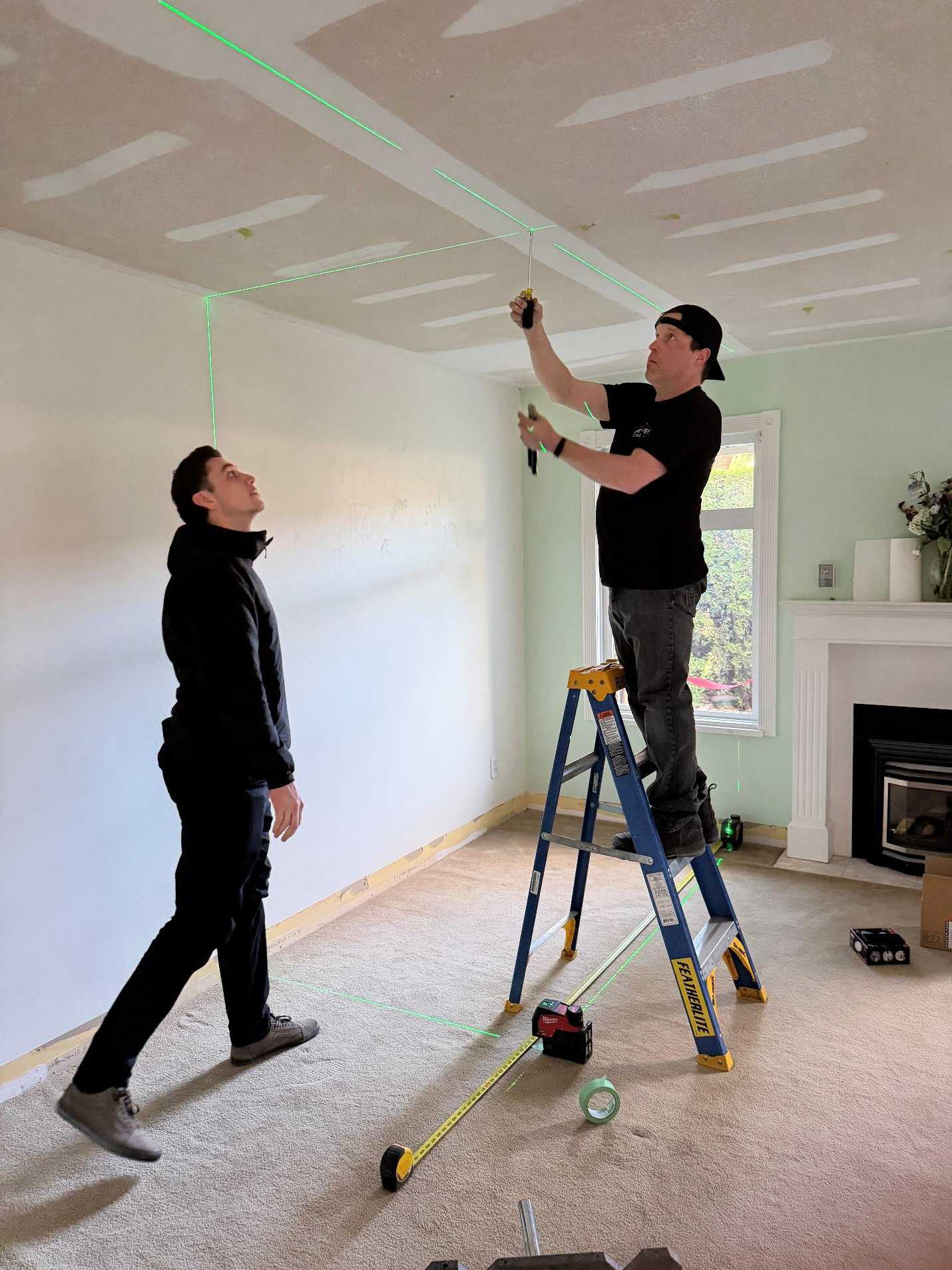 Two electricians using a green laser level and tape measure to ensure perfect alignment for a recessed lighting upgrade in a living room.