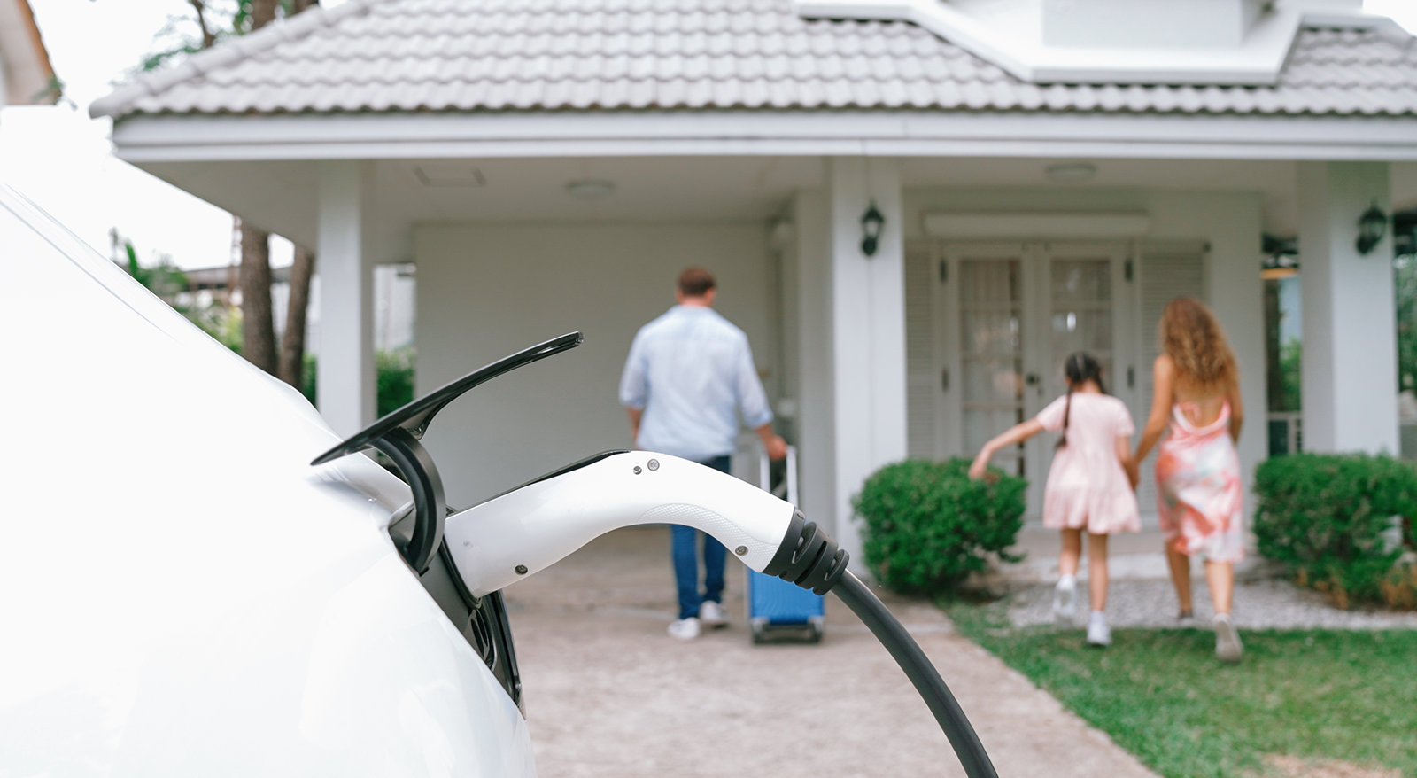Electric vehicle charging plug connected to a car outside a home with a family in the background.